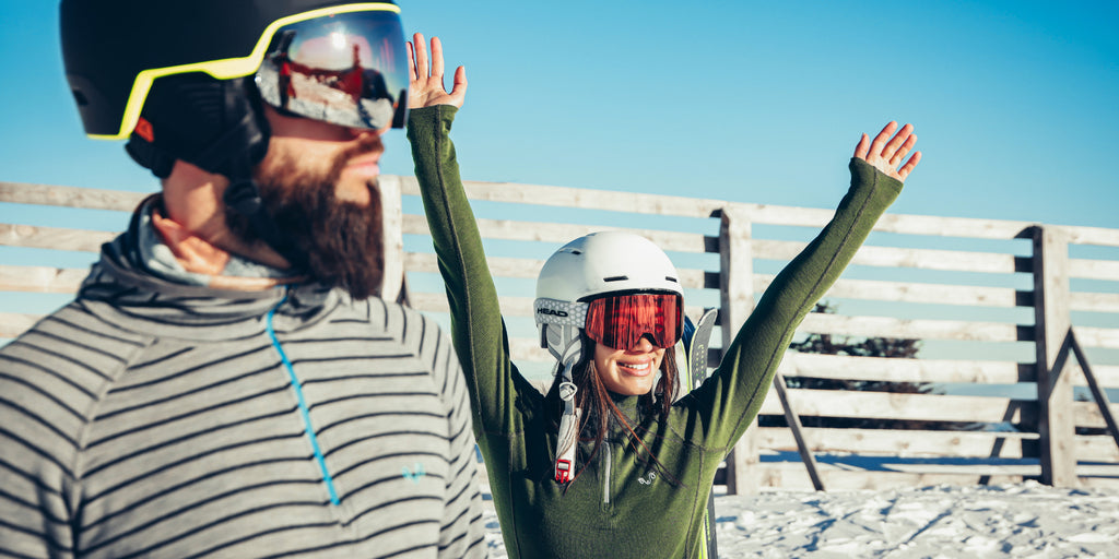 Mann und Frau im Skiurlaub bei Sonnenschein: Frau im grünen Merino-Longsleeve reißt jubelnd die Arme hoch, im Vordergrund steht ein Mann im gestreiften Merino-Shirt. Beide tragen Helm und Skibrille.
