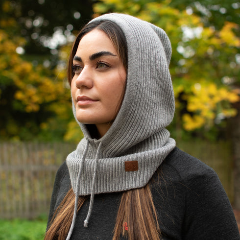Eine Frau in einem oversize Balaclava in der Farbe grau aus Merinowolle, steht in der Natur.