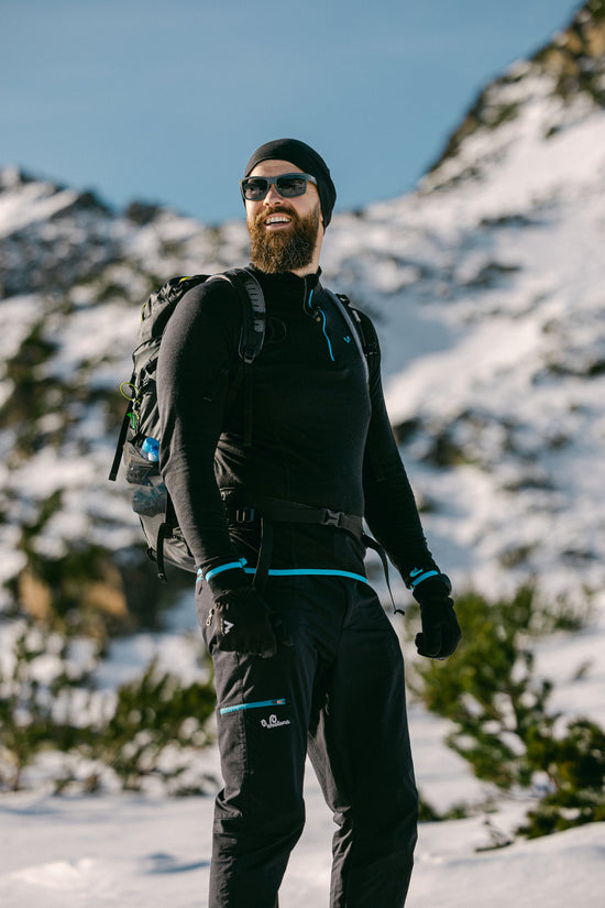 Mann in Merinokleidung von Woolona in den Bergen mit Sonnenbrille, MĂźtze und einem Rucksack auf dem RĂźcken.