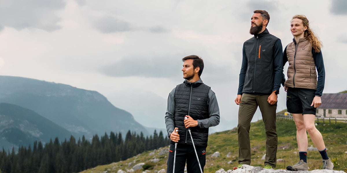 Zwei Männer und eine Frau wandern mit Merinobekleidung in einer bergigen Landschaft. Einer hat Wanderstöcke.