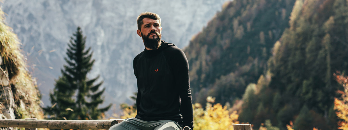 Mann trägt einen Merino-Hoodie und sitzt auf einem Holzbalken in einer bergigen Landschaft im Wald