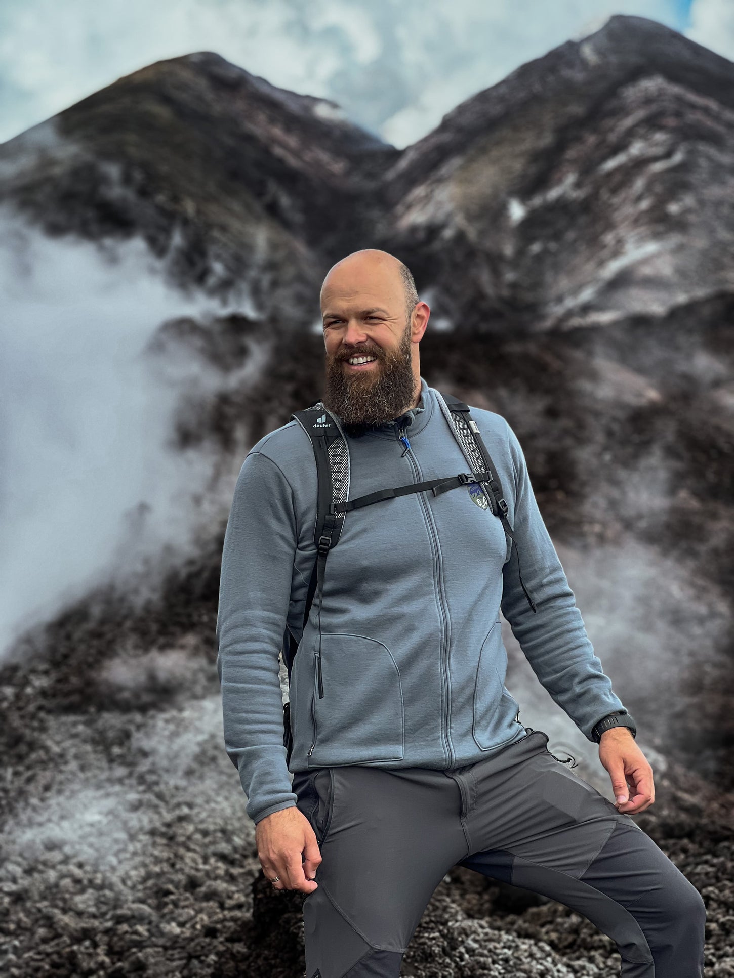 Mann in einer grauen Merino-Jacke mit einem Rucksack steht vor einem schĂśnen Berg