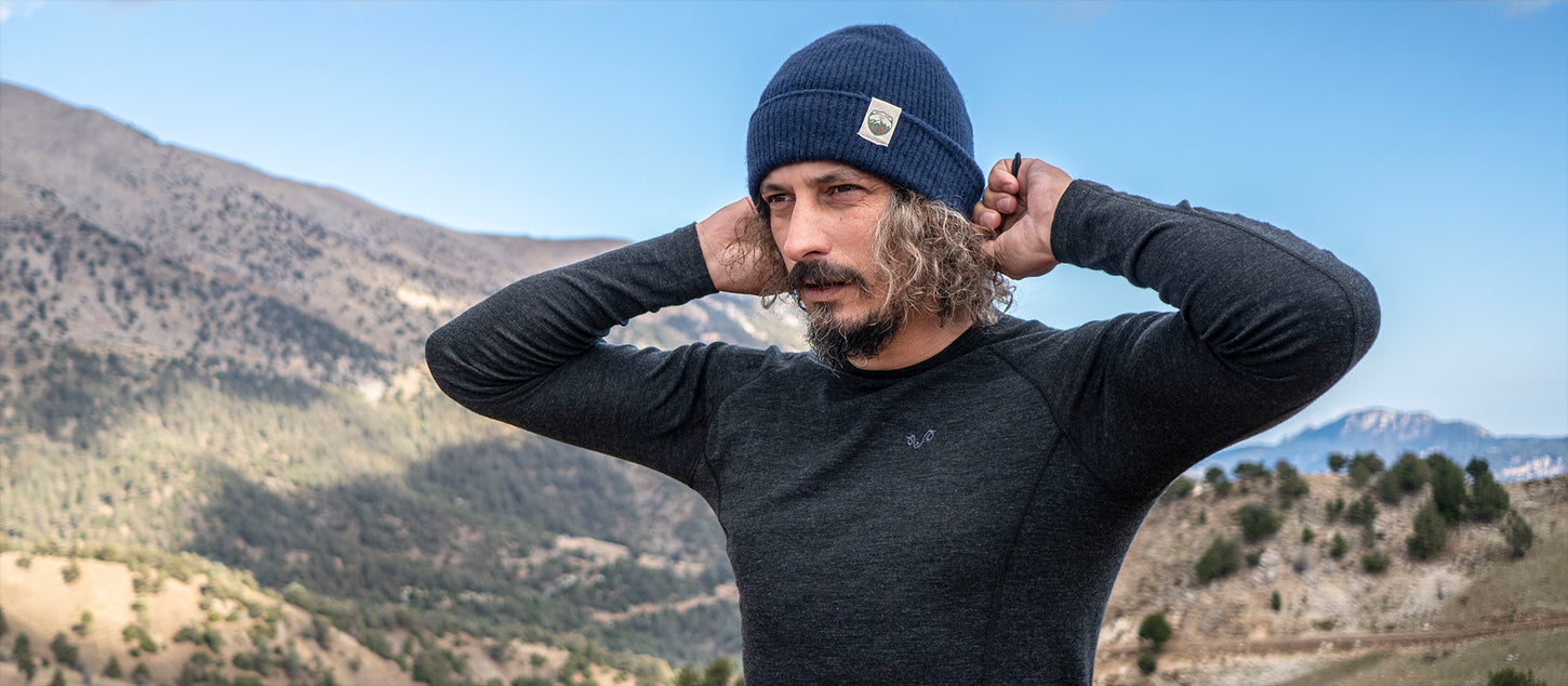 Bärtiger Mann trägt Merino Funktionskleidung und Mßtze und steht in einer bergigen Landschaft