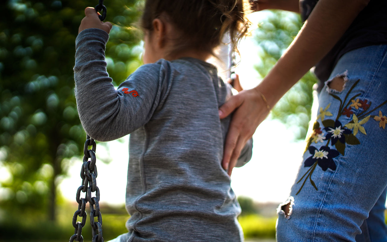 Ein Kind auf der Schaukel trägt Merinokleidung und wird von der Mutter angeschubst