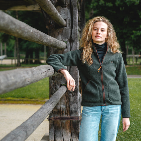 Die Frau lehnt sich an einen Zaun und trägt eine grüne Sherpa Jacke aus Merino und Polyester.