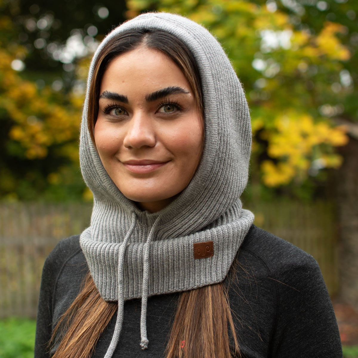 Frau mit einem lächeln, trägt einen grauen Oversize Balaclava Kapuzenschal in der Natur.