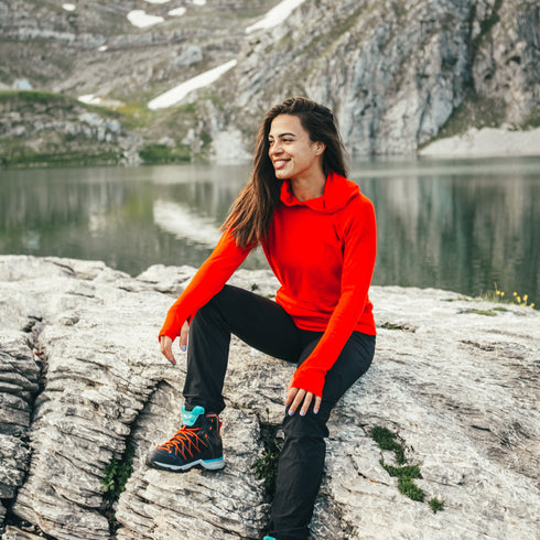 Eine Dame sitzt vor einem See auf einem Felsen und trägt ein rotes langärmliges Merino Kapuzenshirt